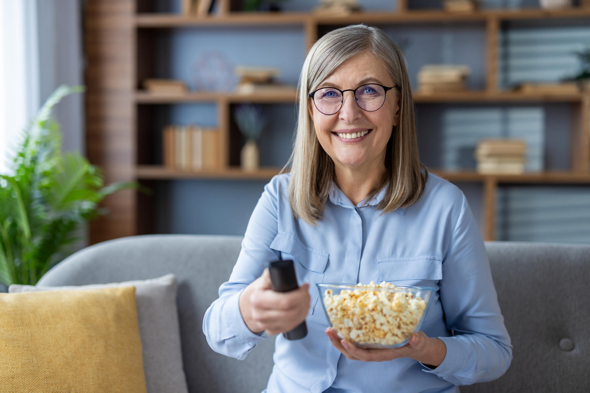Smiling woman holding a bowl of popcorn and remote control while sitting on a cozy sofa. Enjoying leisure time at home with entertainment. Relaxed elderly lady ready to watch TV. Smiling woman holding a bowl of popcorn and remote control while sitting on a cozy sofa. Enjoying leisure time at home with entertainment. Relaxed elderly lady ready to watch TV.