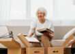 elderly woman sits on a sofa at home with boxes. collecting things with memories albums with photos and photo frames moving to a new place cleaning things and a happy smile. Lifestyle retirement.