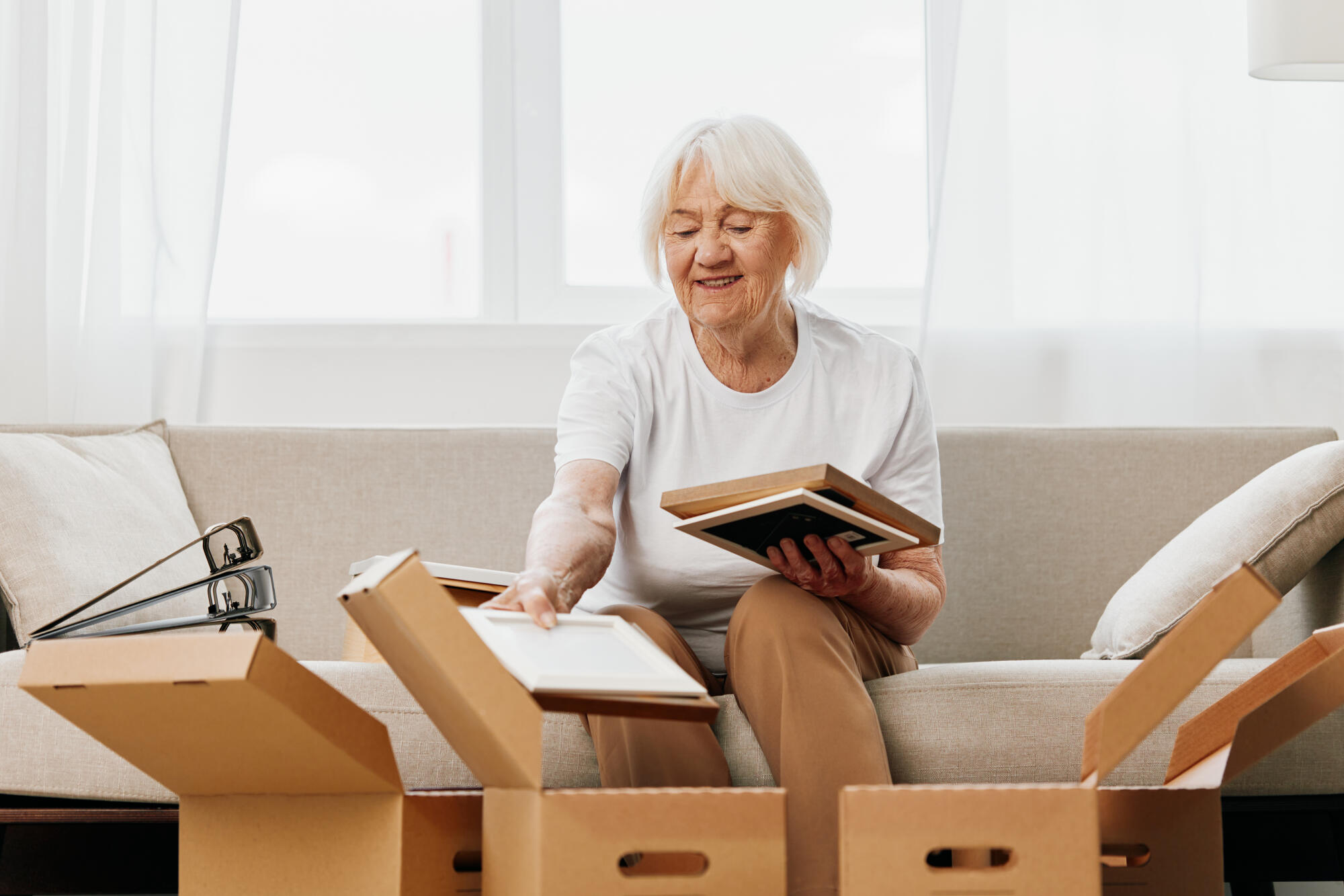 elderly woman sits on a sofa at home with boxes. collecting things with memories albums with photos and photo frames moving to a new place cleaning things and a happy smile. Lifestyle retirement. elderly woman sits on a sofa at home with boxes. collecting things with memories albums with photos and photo frames moving to a new place cleaning things and a happy smile. Lifestyle retirement.