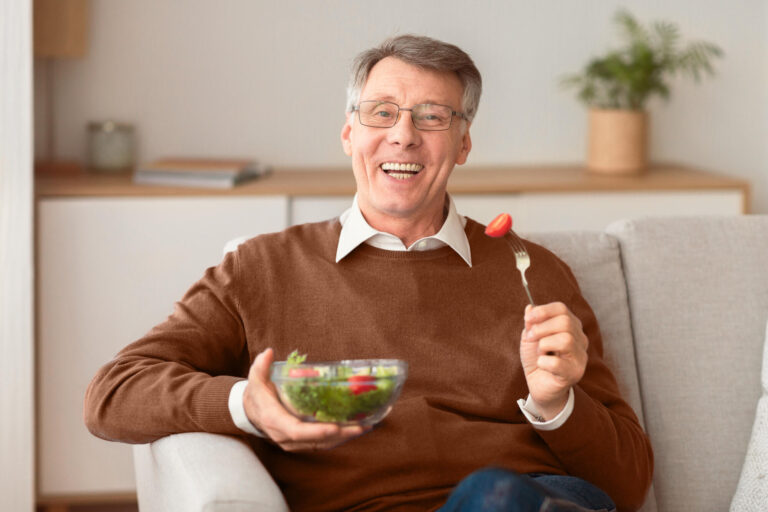 Elderly man eating vegetable salad smiling sitting on sofa