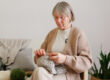 A woman sits in a comfortable chair, focused on knitting a soft gray project. The warm interior has plants and yarn in various colors around her