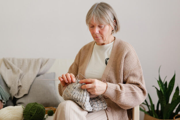 A woman sits in a comfortable chair, focused on knitting a soft gray project. The warm interior has plants and yarn in various colors around her