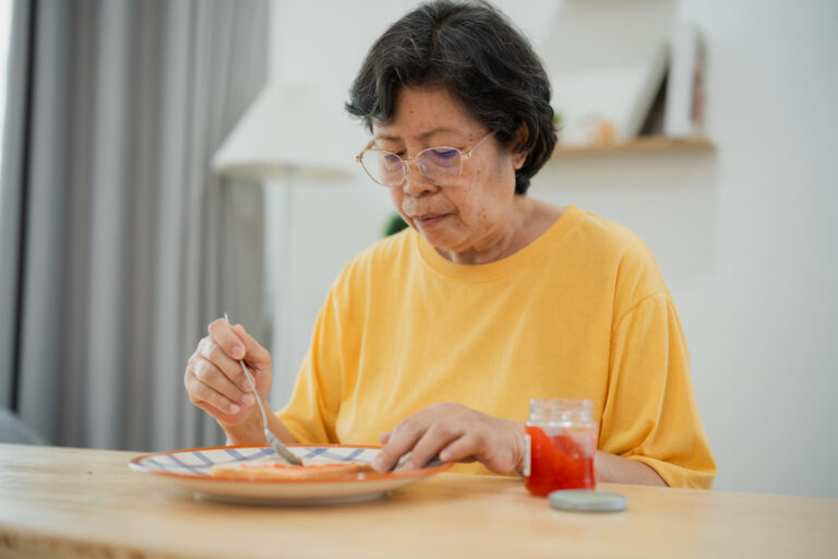 Senior Asian woman enjoying breakfast with toast and jam in cozy kitchen setting, health conscious lifestyle, natural lighting, and simple tableware