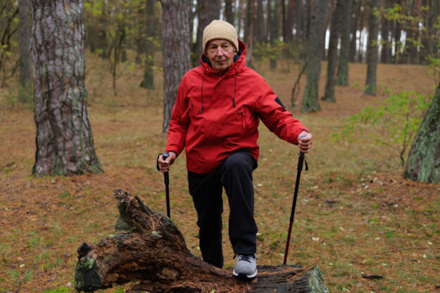 An older man is hiking in a serene forest, wearing a red jacket and a warm hat, enjoying the outdoors while using walking poles for support